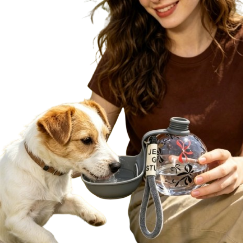 A woman holds a clear water bottle with a gray loop handle while a small white and tan dog drinks from a gray bowl attached to the bottle. The woman is wearing a brown shirt and khaki pants against a plain white background.